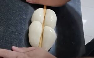 A close-up of a wooden dowel pressing into the center of the twin dough rolls to create the iconic four-segment shape.