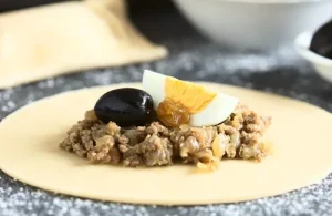 Placing a hard-boiled egg, olives, and pino filling onto a circular dough cutout to assemble the empanada.
