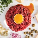 Homemade beef tartare topped with an egg yolk, surrounded by red onion and toast