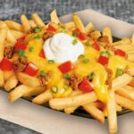 A generous plate of french fries topped with ground beef, melted cheddar cheese sauce, fresh diced tomato, chopped chives, and a touch of plain yogurt.