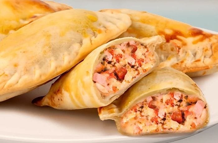 A tray of golden, glossy Chilean Neapolitan empanadas with visible melted cheese, ham, and tomato filling.