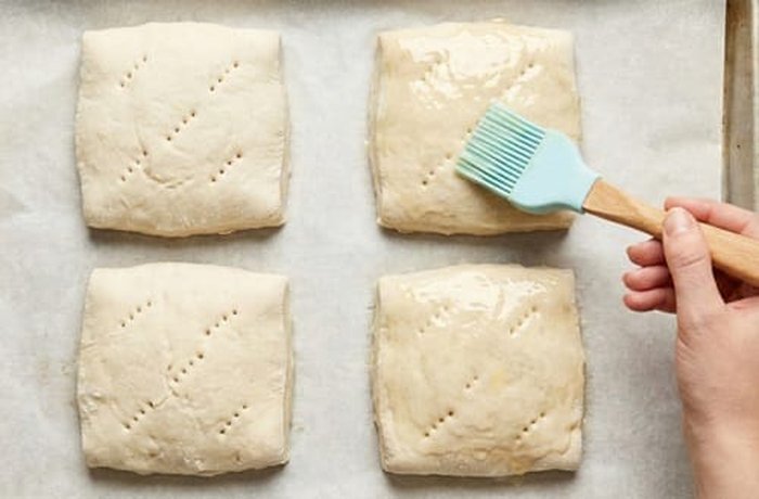 Pan coliza squares on a baking sheet being brushed with egg wash before going into the oven.