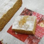 Rectangular Chilean honey bread dusted with powdered sugar, showing a dark spongy crumb, next to a cup of coffee